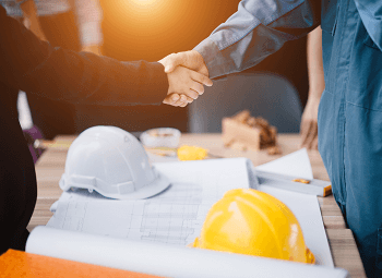 Two contractors shaking hands over a table with plans and hard hats